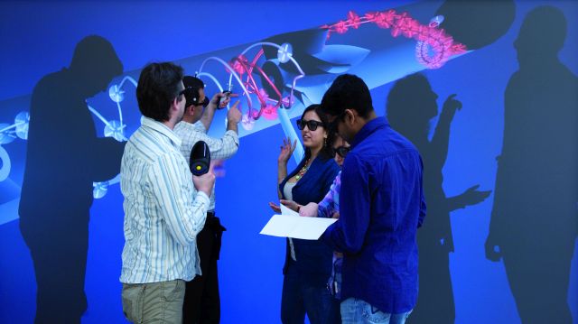 Four researchers are standing in front of a projection of a blood pump, are talking to each other and taking notes and photos.