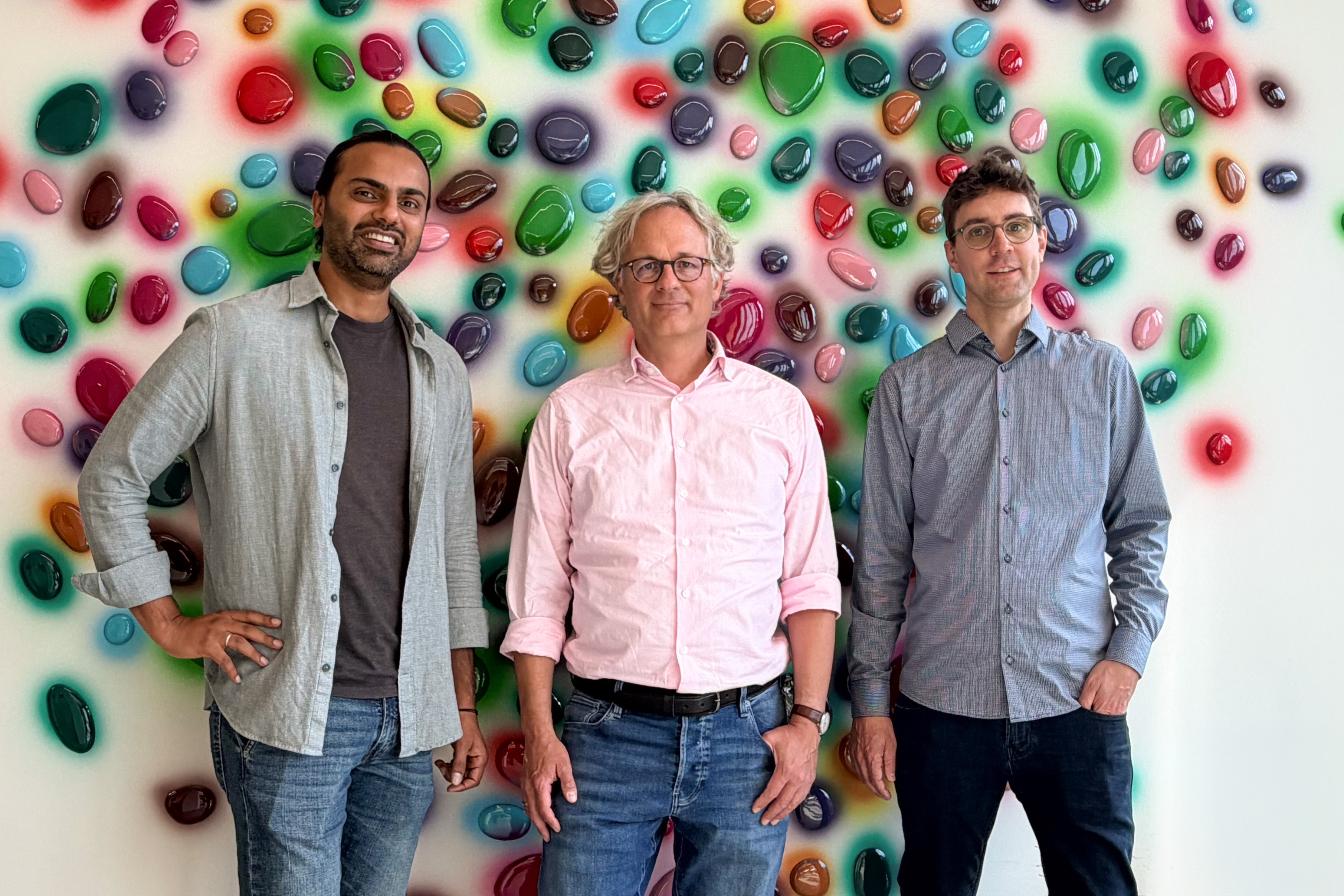 ​​Three men stand side by side in front of a colorful wall decorated with glossy, multicolored pebble-like shapes resembling bacterial colonies.​ 