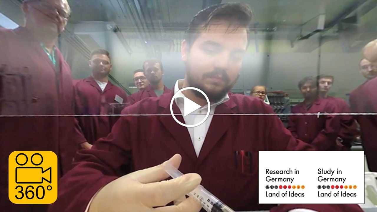 A researcher is working with a syringe. Behind him are standing many other researchers.