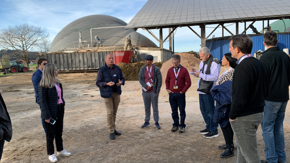 Ten people stand in front of a biogas plant and listen to a presentation by Thomas Pfluger.