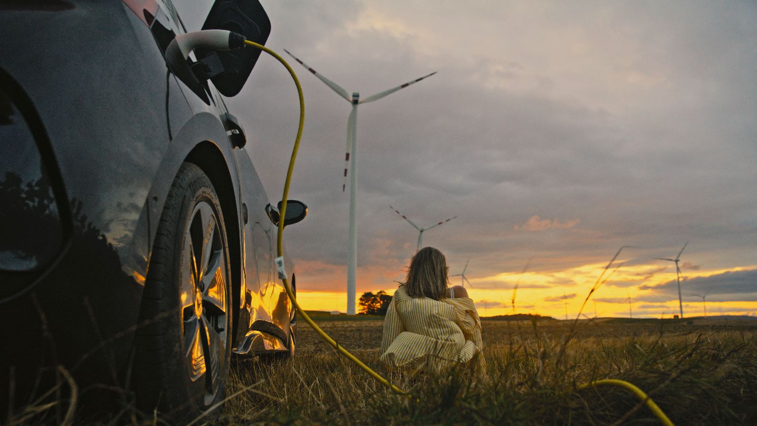 Wide-angle shot of a mid adult woman in casual beige sweater sitting peacefully beside her charging electric vehicle, surrounded by wind turbines against dramatic sunset sky.