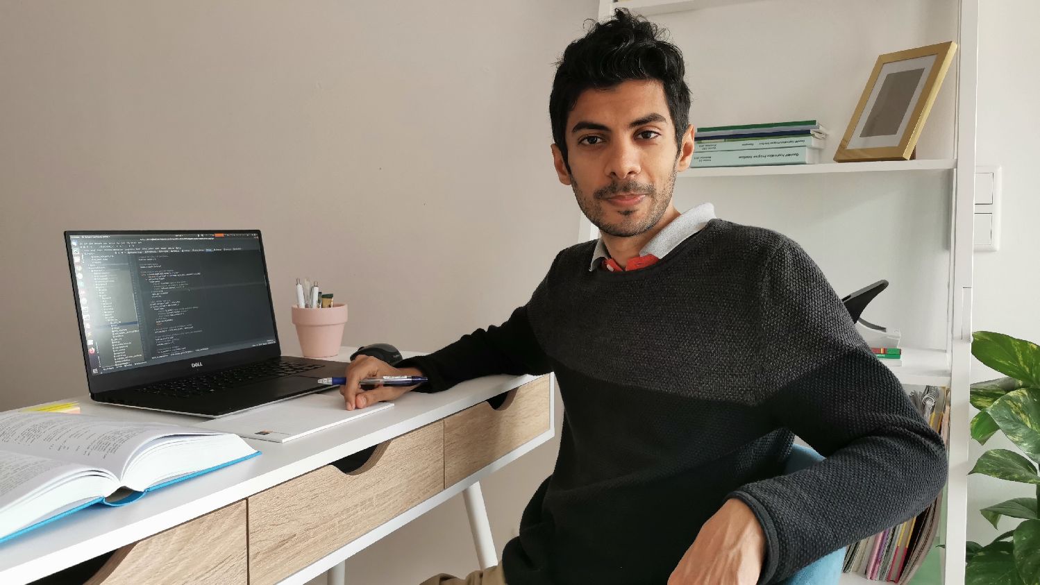 A researcher is sitting at his desk holding a pen in his hand. On the desk are laying his laptop and an open book.