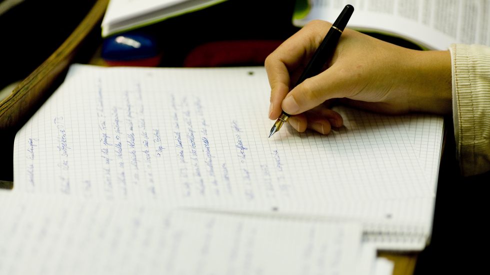 There are several pieces of paper lying on a desk. On the one in focus, there is a hand of a person who is writing on it with a pen.