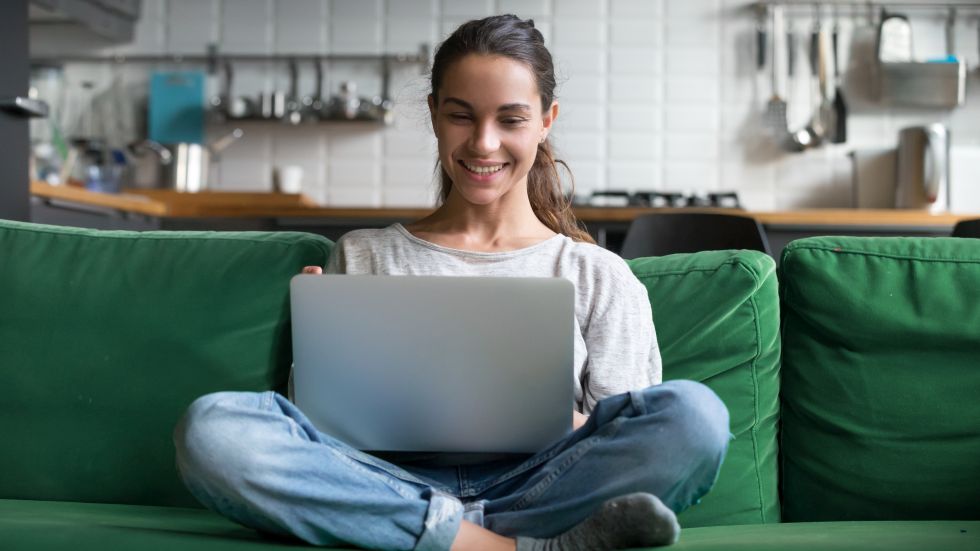 Junge Frau sitzt auf einem grünen Sofa und schreibt lächelnd etwas auf einem Laptop in ihrer Wohnung. 