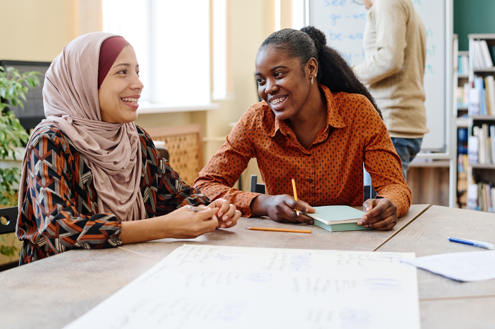 Young black women and young woman from the Middle East sitting at a table and have fun chatting about something during German classes for immigrants.