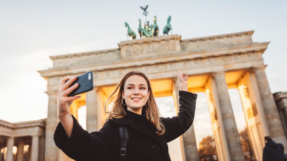 Eine junge, lächelnde Frau steht vor dem Brandenburger Tor, auf das sie mit dem Finger zeigt, und macht dabei ein Selfie. 