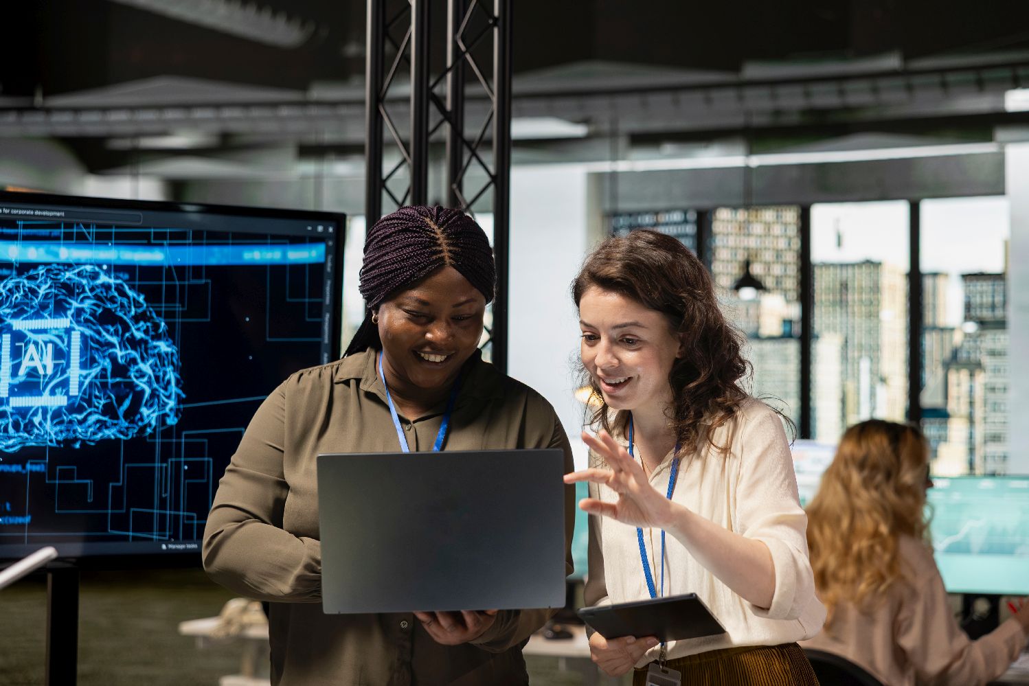 Two young woman excitedly look at laptop held by the left woman. The right woman points at something on the scrren. They appear to be in a modern office environment.