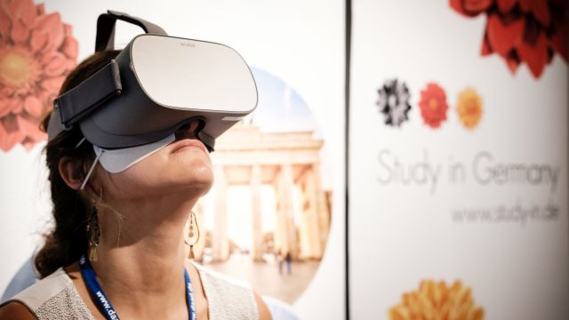 A young women is sitting on a stool in front of a "Research in Germany" banner and looks upwards with virtual reality glasses on her eyes.