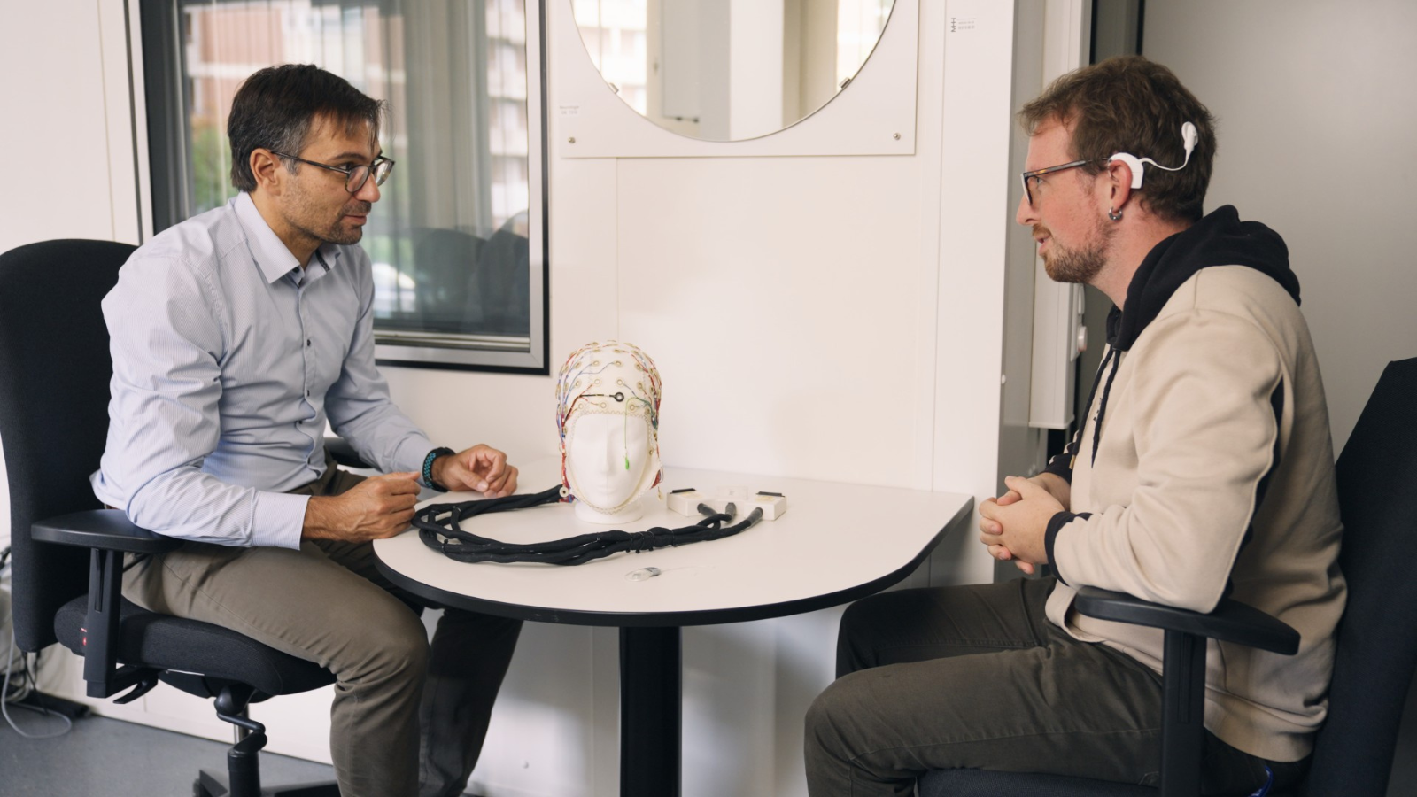 ​​​The picture shows Waldo Nogueira. He talks to a test person who wears a cochlea implant. Between the two men stands a table. On it lie some cables and an EEG cap.​​ 